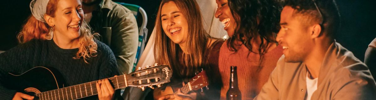 A diverse group of friends playing guitar and ukulele by a campfire at night, having fun and socializing.
